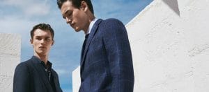 Two male models in navy suits pose outdoors beside white concrete walls against a blue sky spell out a fashion shoot.