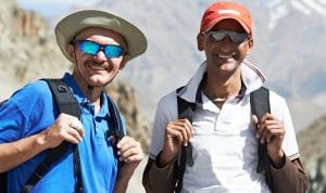 Two men with backpacks posing outdoors in a mountainous landscape, smiling at the camera.
