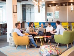 Six coworkers in a modern open-office lounge sit on colorful chairs, chatting with laptops and notebooks on small tables in front of them.