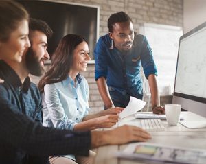 Team of four colleagues collaborating around a desk, looking at a computer monitor on a bright office desk.