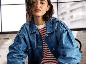 Woman wearing a blue denim jacket over a red striped sweater, posing indoors by large windows.