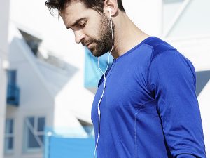 Man in a blue athletic shirt wearing white earbud headphones, outdoors, looking down as he jogs or walks.
