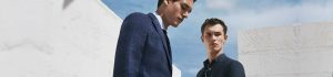 Two men in navy suits stand against white walls outdoors, looking off into the distance in a fashion shoot.