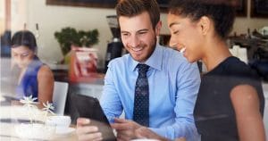 Two colleagues in a cafe looking at a tablet, smiling as they discuss something together.