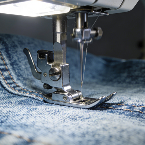 Close-up of a sewing machine presser foot and needle stitching blue denim jeans.
