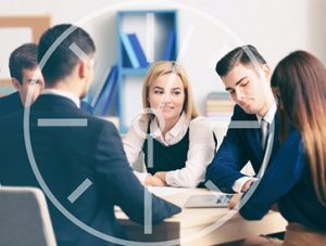 Group of professionals in a meeting around a conference table, with a woman in a white blouse speaking to the team.