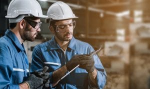 Two factory workers wearing hard hats and safety goggles examine a metal part while holding a clipboard.