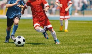 Young soccer players chase a black-and-white ball on a green field, one in red kicking while a blue-uniformed player pursues nearby.