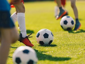 Youth soccer players practice on a sunny green field, with a soccer ball near a player’s red cleats.