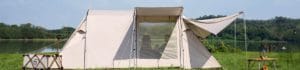 Beige camping tent pitched on grass beside a calm lake with a fabric awning and a window showing people inside.