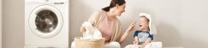 Mother and daughter laugh while folding laundry next to a washing machine, basket of clothes nearby.