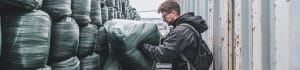 Man with a backpack inspecting large green plastic-wrapped bales in an industrial yard beside metal containers