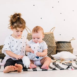 Two young children sit on a striped rug, playing with a small plush bunny toy together in a cozy play area with baskets in the background.