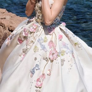 Back view of a person wearing a white floral-embroidered ball gown, standing on a rocky shoreline with the sea in the background at the edge of the water.