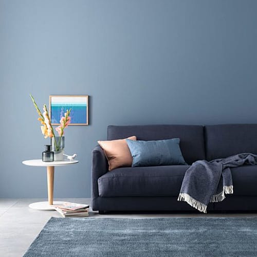 Navy sofa with peach and blue pillows, a fringed gray throw, in a blue-walled living room; white side table with a vase of flowers and framed art on the wall.