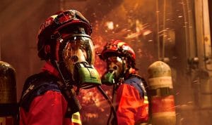 Two firefighters in red turnout gear with full-face respirators stand in a smoke-filled scene, sparks flying as they work.