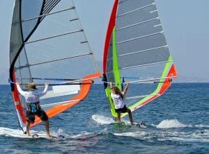 Two windsurfers ride side by side on blue ocean water with bright sails catching wind.
