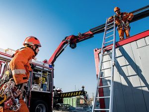 Firefighters in orange gear operate a crane arm and ladder at a construction site, coordinating a high-point task under a clear blue sky.