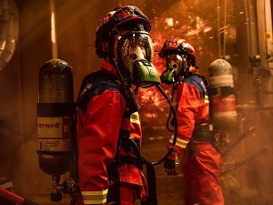 Two firefighters in red turnout gear with helmets and air tanks move through a smoky, fiery scene.