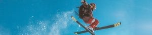 Skier in red pants wearing a helmet and goggles flies mid-air with skis crossed, powder snow spraying against a clear blue sky carrying out a freestyle jump.