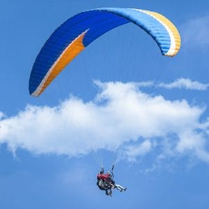 Two people paragliding under a blue-and-yellow wing against a bright sky, seated in harnesses with cords connecting to the canopy.