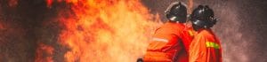 Two firefighters in orange turnout gear and helmets battle a large blaze with water spray surrounding them.