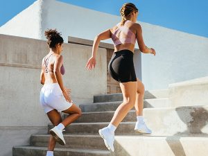 Two women in athletic outfits running up outdoor concrete stairs on a sunny day.
