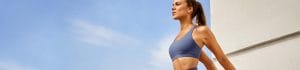 Fit woman in a blue sports bra standing outdoors against a clear blue sky and pale wall.