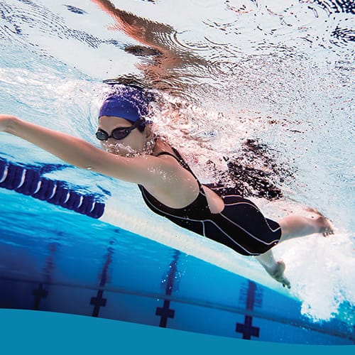 Swimmer in a black swimsuit and blue cap swims freestyle underwater in a blue pool with goggles on.