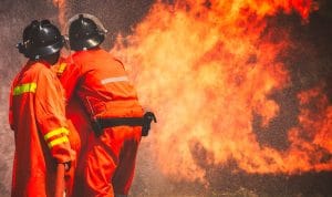 Two firefighters in bright orange gear fight a intense blaze, flames roaring nearby.
