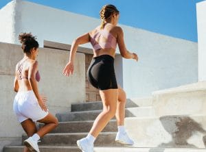 Two women in athletic wear run up concrete outdoor stairs on a sunny day, blue sky above, with white walls nearby.
