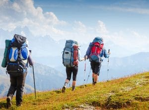 Three hikers with large backpacks trekking along a mountain trail, with snowy peaks in the distance and blue sky above.
