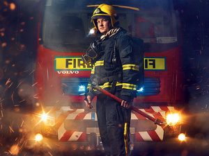 Firefighter in full turnout gear stands in front of a red fire engine, holding a tool amid sparks and emergency lights.