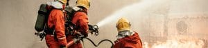 Firefighters in orange protective gear spraying water from a hose at a blaze