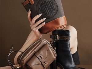 Close-up of fashion bags and a heeled boot arranged together: tan crossbody, brown leather pouch, and black ankle boot against a brown background.