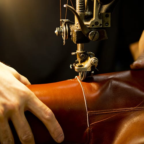 Close-up of a sewing machine needle and presser foot stitching brown leather, with hands guiding the material.