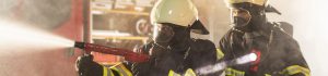 Two firefighters in protective gear aim a red hose and spray water during a training exercise in a smoky environment, with one watching ahead.