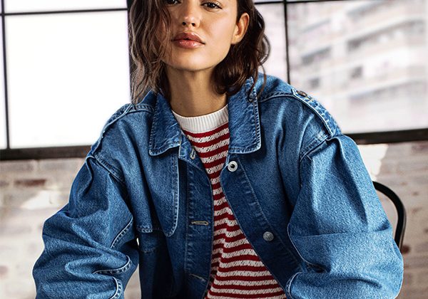 Woman wearing a blue denim jacket over a red striped sweater, posing indoors by large windows.