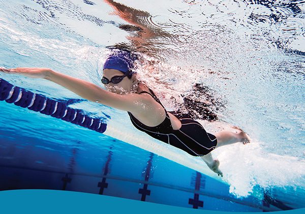 Swimmer in a black swimsuit and blue cap glides freestyle in a pool, wearing goggles beside a lane rope.