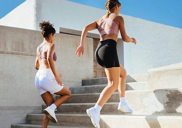 Two women in athletic outfits running up outdoor concrete stairs on a sunny day.