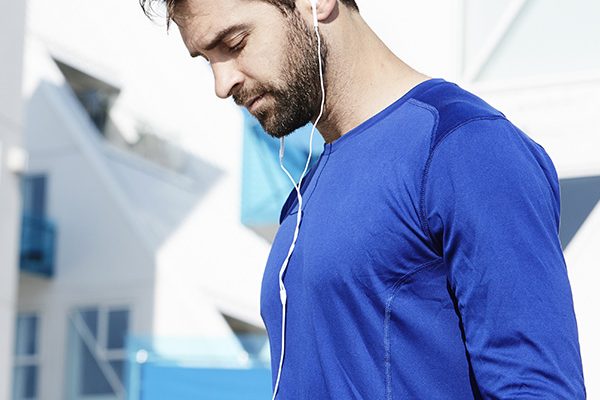Man in a blue athletic shirt wearing white earbud headphones, outdoors, looking down as he jogs or walks.