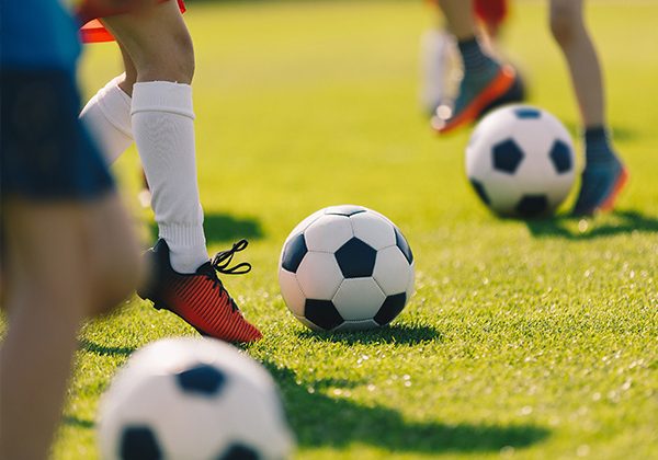 Youth soccer players practice on a sunny green field, with a soccer ball near a player’s red cleats.
