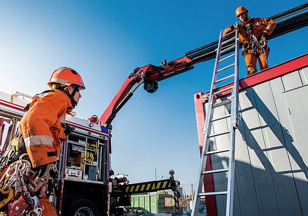 Firefighters in orange gear operate a crane arm and ladder at a construction site, coordinating a high-point task under a clear blue sky.