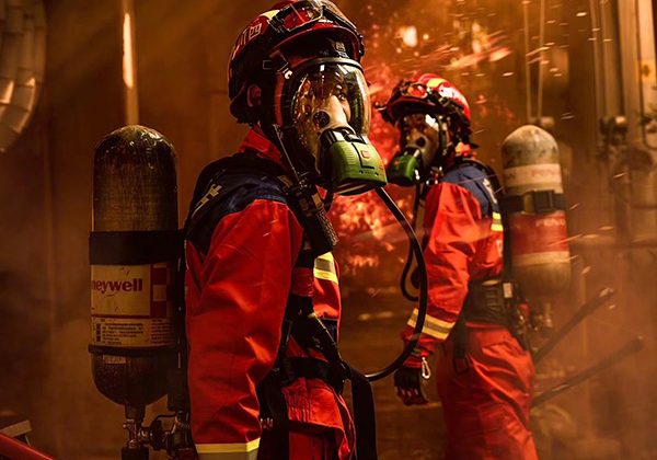Two firefighters in red turnout gear with helmets and air tanks move through a smoky, fiery scene.