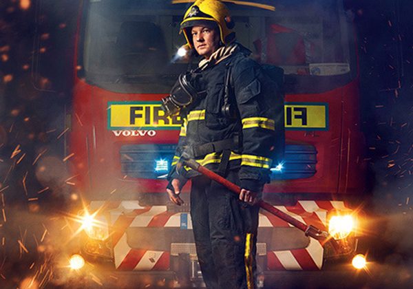 Firefighter in full turnout gear stands in front of a red fire engine, holding a tool amid sparks and emergency lights.