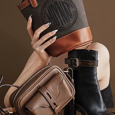Close-up of fashion bags and a heeled boot arranged together: tan crossbody, brown leather pouch, and black ankle boot against a brown background.